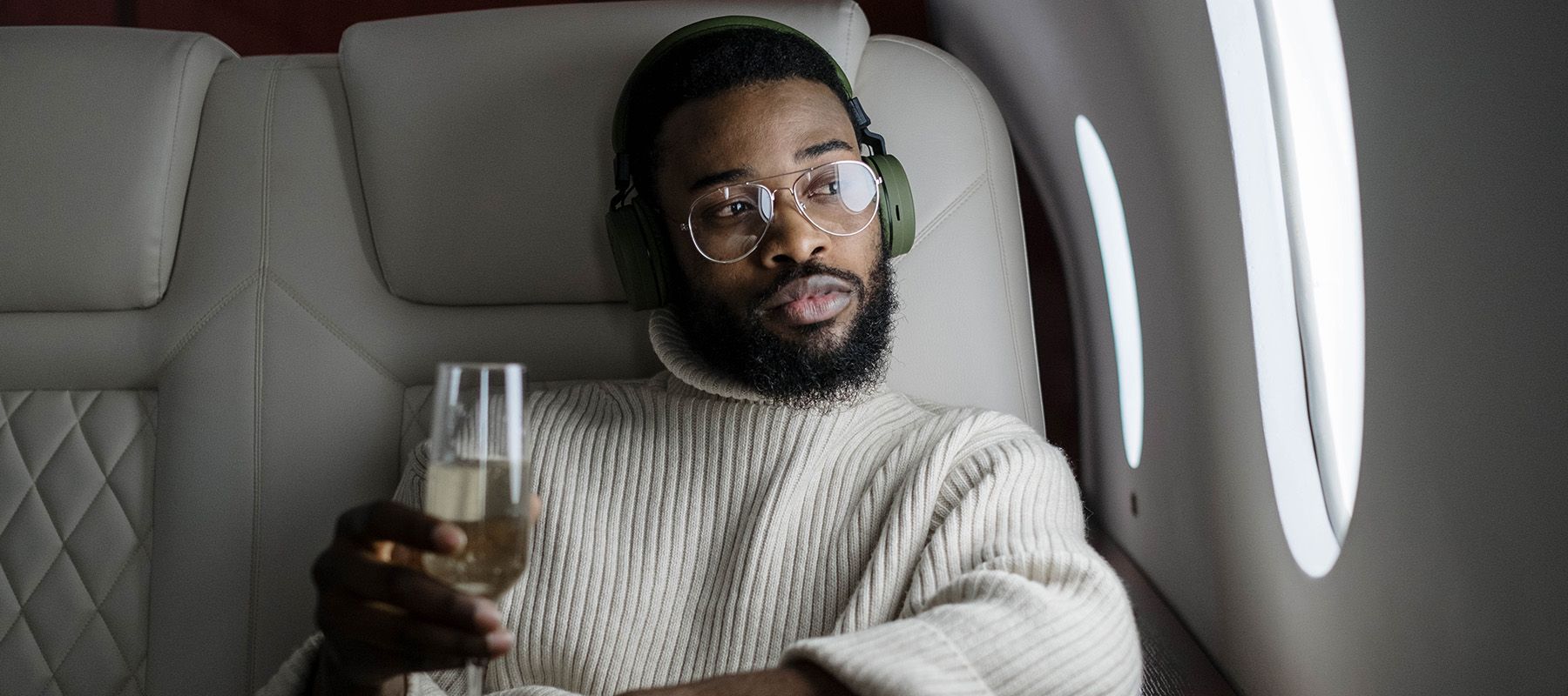 Black man sitting in first-class on plane wearing headphones looking out window holding champagne glass